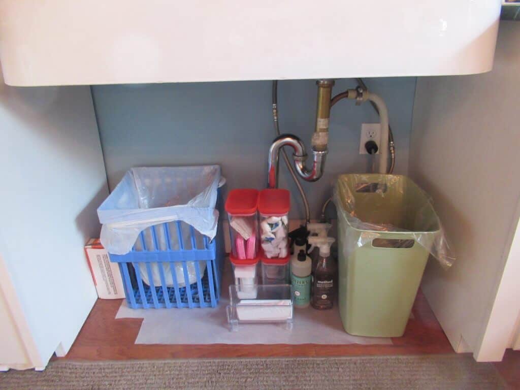 Under kitchen sink blue recycle bin, pipes, various cleaning supplies, and green trash can