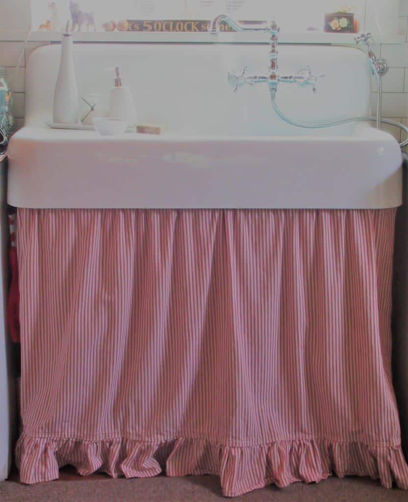 White, antique-style farmhouse sink with red and white ticking stripe curtain; chrome faucet, white and chrome soap dispensers